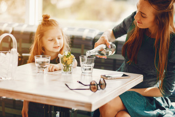 young and stylish mother with long hair and a green dress sitting with her little cute daughter in the summer cafe and pours water into a glass