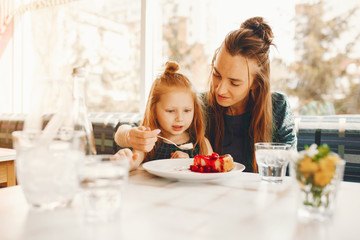 young and stylish mother with long hair and a green dress sitting with her little cute daughter in the summer cafe and she feeds her daughter with a dessert