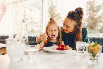 Obraz na płótnie Canvas young and stylish mother with long hair and a green dress sitting with her little cute daughter in the summer cafe and she feeds her daughter with a dessert