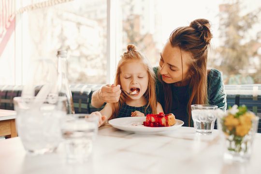 young and stylish mother with long hair and a green dress sitting with her little cute daughter in the summer cafe and she feeds her daughter with a dessert