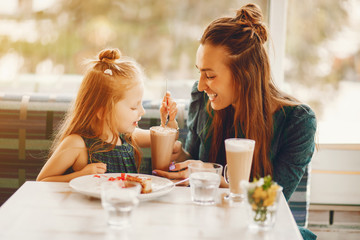 young and stylish mother with long hair and a green dress sitting with her little cute daughter in the summer cafe and she feeds her daughter with a dessert