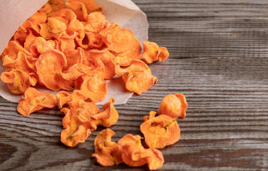 Close-up orange healthy carrot chips falling from white paper bag to old wooden table. 