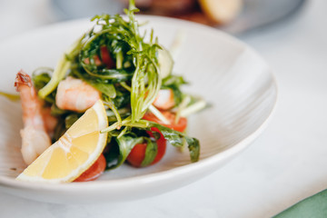Closeup Salad with shrimps, arugula, lemon, tomatoes in deep bowl. Table setting is light