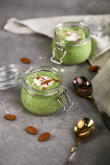Two glass jars full of green smoothie with yogurt and almonds on grey table with metal spoons and linen canvas