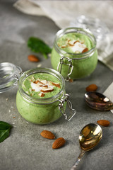 Glass jars full of green smoothie with almonds and yogurt on grey table with linen canvas and metal spoons
