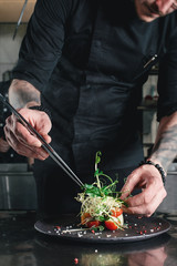 Chef finishing healthy salad on a black plate with tweezers. almost ready to serve it on a table