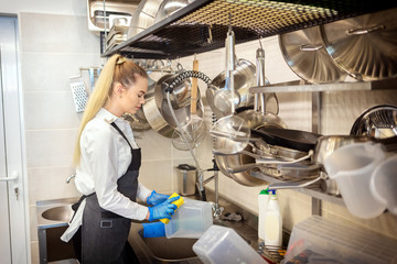 Young woman washing dishes using sponge in restaurant sink at end of working day 