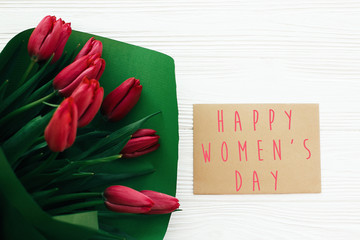Happy Women's Day text and beautiful red tulips on white wooden background flat lay. Happy women day greeting card with spring flowers. Stylish simple holiday card