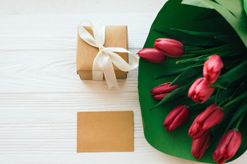 Beautiful red tulips bouquet in green paper,gift box and craft card with space for text on white wooden background flat lay. Happy mothers day. Pink tulips on white wood. Greeting card template