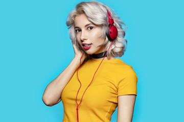 Portrait of adorable pretty light-haired model looking at camera and posing in studio wearing headphones. Stylish lady in yellow shirt. Lifestyle and music concept. Isolated on blue