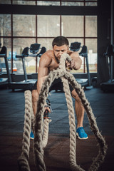 Obraz na płótnie Canvas Good looking young man with beard screaming doing Battle of the ropes with crossfit rope in the hand at the gym opposite the camera. Beginner's Guide to Battling Ropes. Crossfit Rope Training