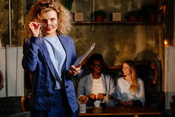 Couple consultation. Happy pretty professional blond curly female psychologist wearing glasses smiling to the camera and carrying pencil and folder. Multiethnic couple on background