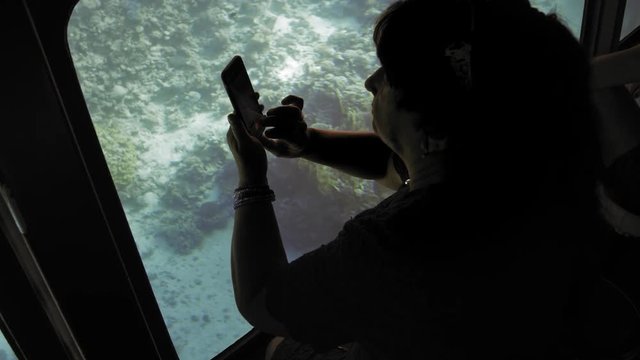 Senior female adult tourist on board a submarine for viewing coral reefs of the seabed photographs fish on a smartphone's chamber. Slow Motion.