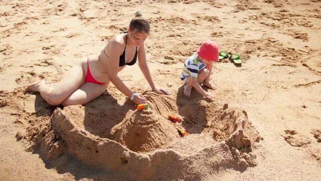 4k footage of young mother in bikini building sand castle on the sea beach with her 3 years old son