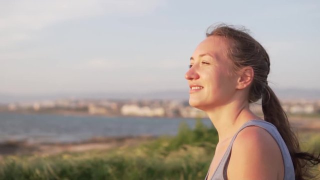 Beautiful young woman enjoying the view of the sea and nature from a high cliff. She smiles and squints from the bright sun. Her hair develops on the sides of the wind. girl puts her hand to her face