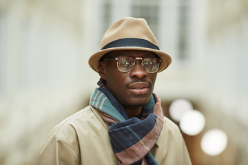 Head and shoulders portrait of trendy African man wearing hat and trenchcoat looking at camera while posing in beautiful building outdoors, copy space