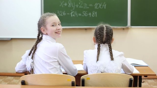 Schoolgirl girls look at the board view from the back Schoolgirls teenagers sitting at the table watching the school board on ocher turn back look back. Girls in white blouses, hair braided in braids