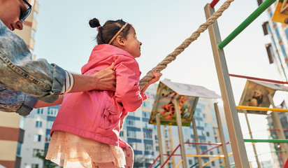 Side view image of active cute little girl climbing on a rope at playground. Mother helping her daughter to climb on a wall with a rope outside. Motherhood and childhood concept. Lifestyle, education.