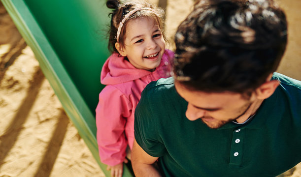 Top view image of cute happy kid daughter playing with her father outdoors. Handsome dad and toddler little girl laughing and having fun and playing at slide playground. Father and daughter share love