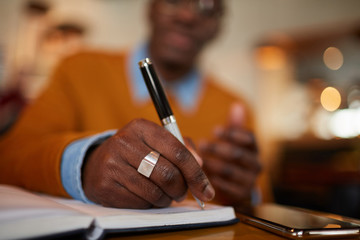 Close up of contemporary African man holding pen while working  sitting at table in cafe, copy space