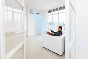 Young Man On Sofa Operating Air Conditioner With Remote Control