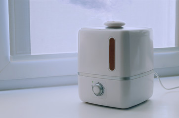 Humidifier in the dwelling. A small white portable device to create a comfortable humidity in the space of the apartment throws a stream of steam. Against the background of a window in the afternoon.