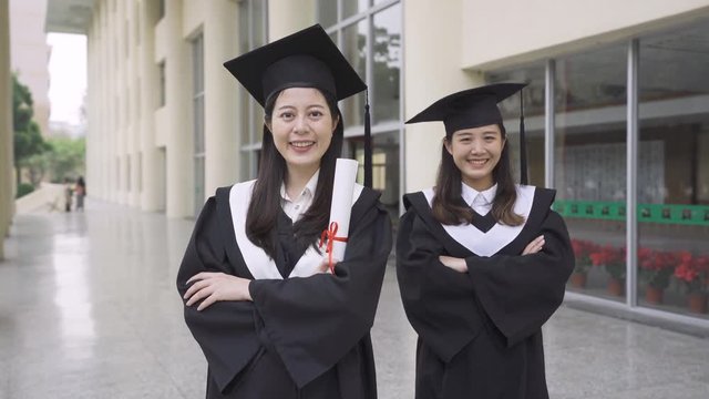 two excited happy asian female students standing in university hall in mantle with diploma in hand smiling and face looking at camera. confident fresh graduate crossed arms attractive smile.