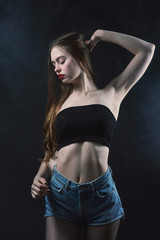 Fototapeta na wymiar Young beautiful, slender, passionate brown-haired woman with bright red lips and long hair in a black top and denim shorts on a dark background