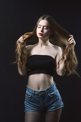 Young beautiful, slender, passionate brown-haired woman with bright red lips and long hair in a black top and denim shorts on a dark background