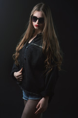 Young beautiful, slender, passionate brown-haired woman with bright red lips and long hair in a black top and denim shorts on a dark background
