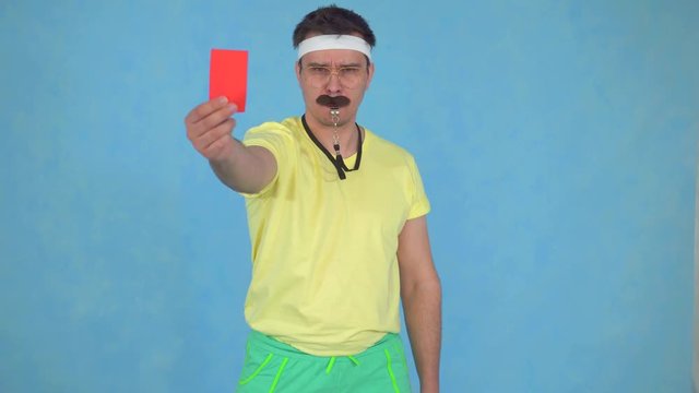Funny disgruntled man coach with whistle from 80's shows red card on blue background