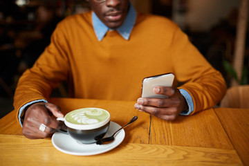 Warm toned shot of contemporary African-American man using smartphone while sitting at table in coffee shop, copy space