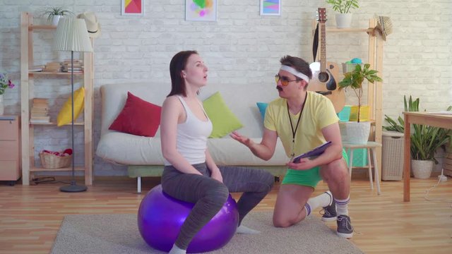 Funny coach man with a mustache from the 80s trains a girl on a fitness ball in his house