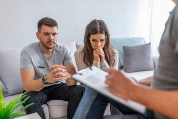 Cheerless upset young woman covering her face and crying while sitting together with her husband at the psychologists office. Unhappy conflicted couple consulting marriage problems with counselor