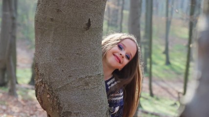 little barefoot girl runs in a forest or Park and hides behind trees in a white dress with blue patterns. - Powered by Adobe