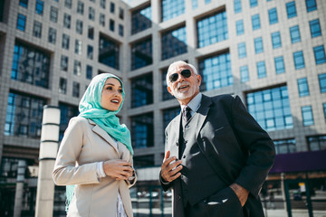 Young beautiful muslim woman with hijab standing in front of modern city building and talking with senior business man. Global investment business concept.