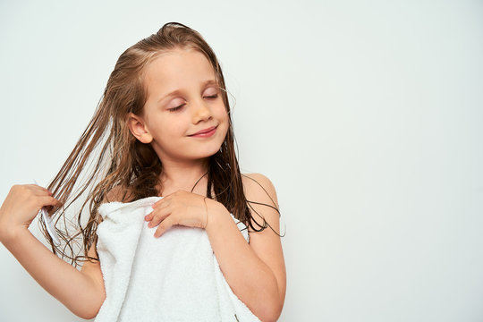 Smiling little preschool girl with wet hair photographed against white background wrapped in white towel while brushing her hair by drawing a comb through wet hair