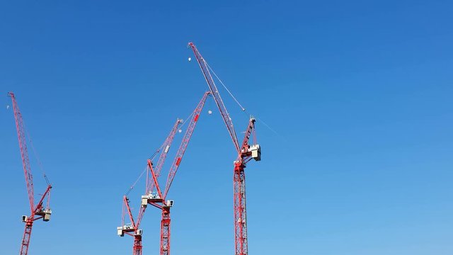 Three construction cranes of red color which stand near against the blue cloudless sky. The view from the pleasure boat