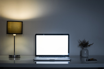 Cozy home office desk at night lit by a lamp and white laptop screen. Clean modern workplace for telework or freelance activity, low key mood