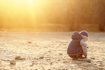 A six-year-old boy with autism sits on the ground alone at sunset. Lonely little child playing with stones outside. The concept of autism and loneliness. Copy space for your text. Setting sun.