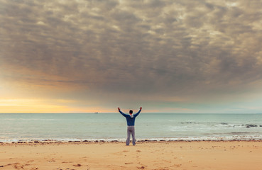 Caucasian man standing at the ocean water on beach at sunset and raised hand