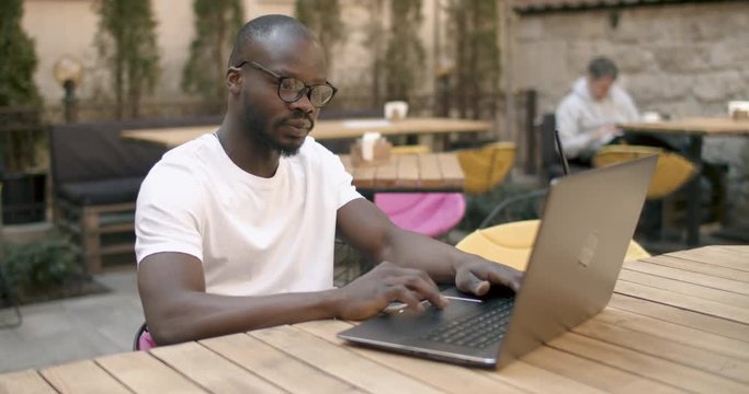 Afroamerican student dressed in white t-shirt opening laptop in outdoors cafe