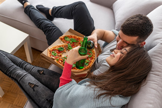 Happy couple drinking beer and eating pizza, spending time together at home. Happy young couple preparing pizza in their home. Beautiful young couple in casual clothes are eating pizza, drinking