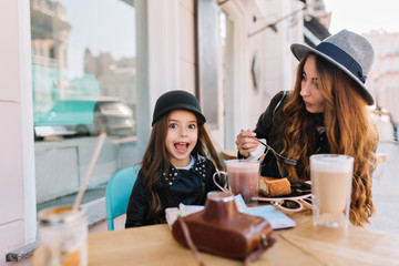 Amazing young woman in vintage gray hat eating tasty cheesecake with her little sister in weekend. Surprised brunette girl enjoying milk shake in outdoor cafe, while her pretty mom drinks coffee