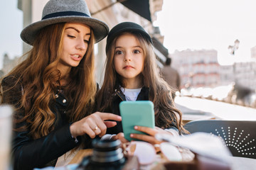 Astonished long-haired little girl holding blue smartphone, while her mom pointing with finger at something important. Portrait of stylish woman in felt gray hat and her charming daughter in cafe