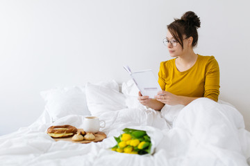 Young woman reading a book and drinking coffee in bed