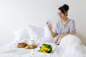 Young woman having breakfast in bed and using mobile phone