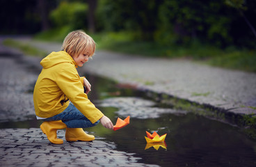 young boy in rain boots and coat is putting paper boats on the water, at spring rainy day