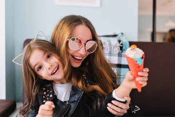 Trendy laughing mom in sunglasses having fun with excited daughter indoors. Portrait of cute...