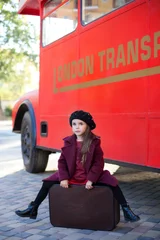 Foto op Plexiglas Little cheerful girl near the red English bus in a beautiful coat and with a suitcase. A little girl is sitting on a suitcase near the bus. London red bus. Spring. Autumn. Journey. London, England. © stock_studio